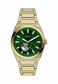 Montre-bracelet en métal doré avec cadran texturé vert, index dorés, aiguilles, petit cadran des secondes et mouvement mécanique interne visible.