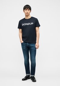 Jeune homme debout les mains dans les poches, portant un t-shirt noir "DONDUP", un jean bleu foncé avec les revers retroussés et des chaussures noires sur fond blanc.