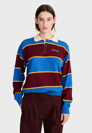 Femme portant un maillot de rugby rayé bleu et bordeaux avec des lignes jaunes et un pantalon marron, debout devant un fond clair uni.