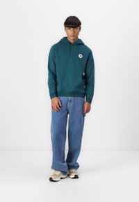 Teal hettegenser med frontlomme og logo på brystet, kombinert med løse blå jeans og nøytrale sneakers på en ensfarget bakgrunn.