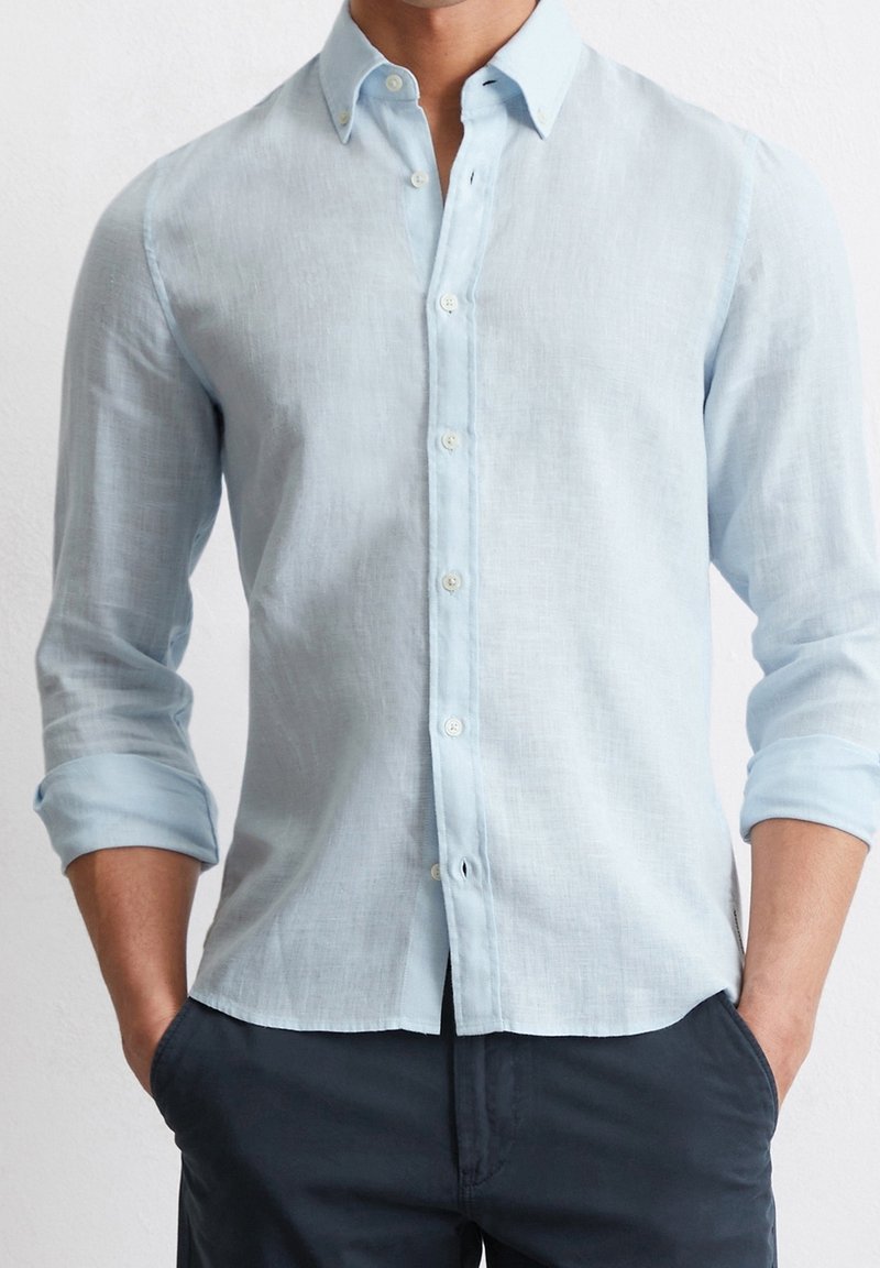 Uomo che indossa una camicia a maniche lunghe azzurro chiaro con le maniche arrotolate e pantaloni scuri, con le mani in tasca, su uno sfondo bianco.