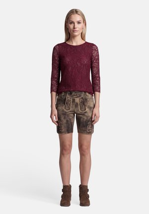 Vrouw staand, gekleed in een bordeauxrode kanten top met driekwart mouwen, bruine leren shorts met borduursel en bruine enkellaarzen.
