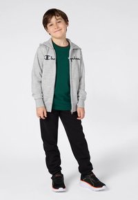 Šedá mikina na zip s logem "Champion", zelené tričko, černé kalhoty a černé tenisky s oranžovými detaily; ležérní sportovní outfit.