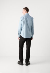 Camisa de manga larga azul claro con cuello abotonado, combinada con pantalones negros y zapatos negros, vista desde atrás contra un fondo liso.