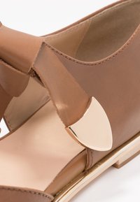 Ruske nahkalevye sandaalit, joissa on leveä hihna, kultaiset yksityiskohdat ja matala pohja, jossa on hieno kiilto. Sisäpuoli on vaaleanbeige.