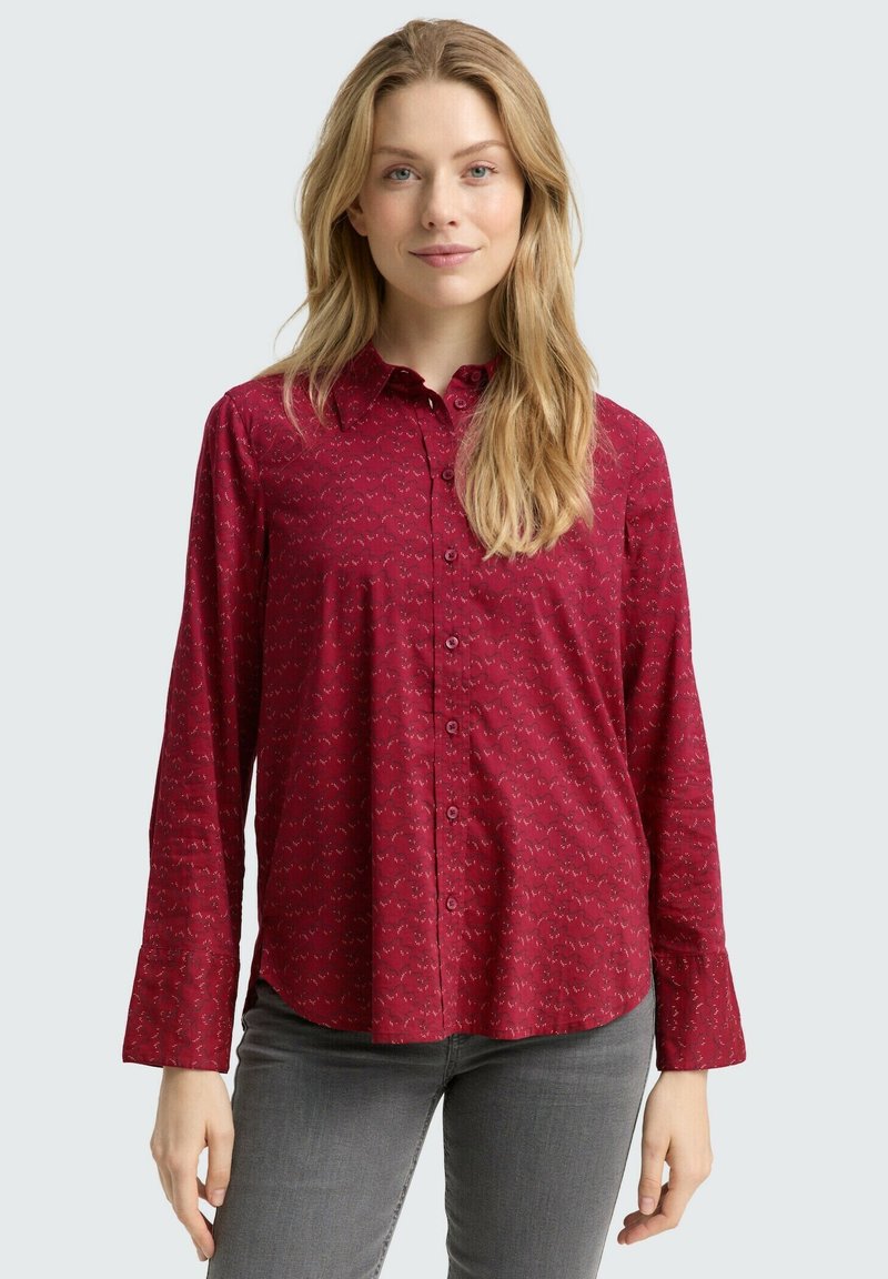 Rotes Hemd mit Knöpfen und dezentem Muster, lange Ärmel und Kragen. Getragen zu grauen Hosen mit glatter Stofftextur.