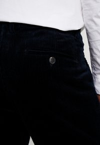 Tmavě námořnické kordové kalhoty s vertikálním žebrováním, jedním knoflíkem a boční kapsou pro praktické použití.