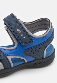 Blå och grå sandal med andningsbart nät, förstärkta remmar och justerbar Velcro-stängning. Har perforerade detaljer och en texturerad sula.