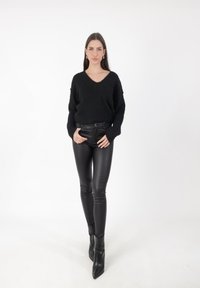 Maglione nero a maglia con scollo a V, abbinato a pantaloni skinny in finta pelle nera e stivaletti neri a punta, su uno sfondo bianco.