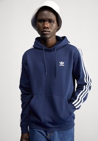 Mörkblå hoodie med vit Adidas-logotyp och tre vertikala vita ränder på ärmarna. Har en känguruficka och justerbar dragsko i huvan.