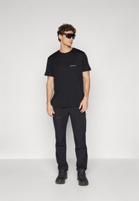 Black t-shirt with subtle blue "icebreaker" logo, paired with black cargo pants and black hiking shoes, worn by a male model with curly hair.