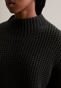 Schwarzer Strickpullover mit einem hohen, gerippten Kragen. Die Textur ist dicht und gemustert, mit einem strukturierten, taillierten Design.