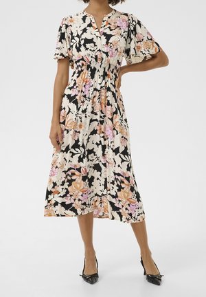 Femme portant une robe midi fleurie à manches courtes volantées et des talons noirs à bouts pointus, debout avec une main sur la hanche.