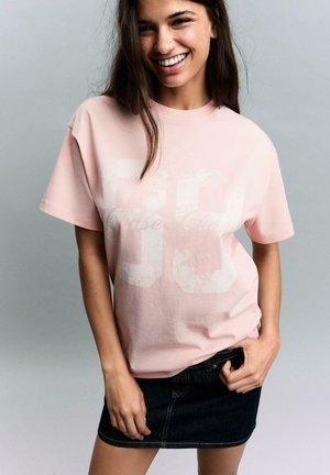 Ung kvinde smilende, iført en lyserød oversized t-shirt med falmet hvidt tryk og en mørk denim mininederdel, stående mod en grå baggrund.