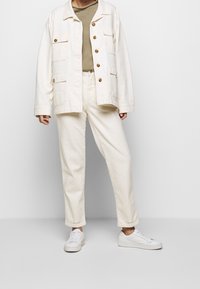 Biała kurtka z płótna z dużymi kieszeniami, beżowy sweter, ecru spodnie i białe trampki, wszystko w luźnym kroju. Gładka tekstura, minimalistyczny design.