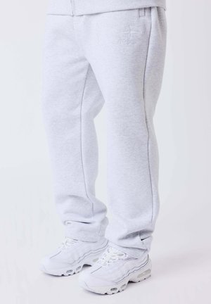 Pantalon de survêtement gris clair avec le logo PXG sur la cuisse, associé à des baskets blanches sur un fond clair uni.