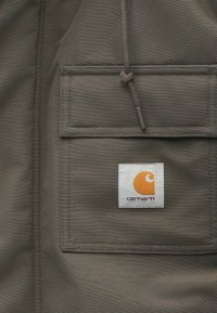 Giacca in tessuto grigio con una tasca frontale e un logo quadrato bianco. Il logo include un vortice arancione e il testo "carhartt" qui sotto.