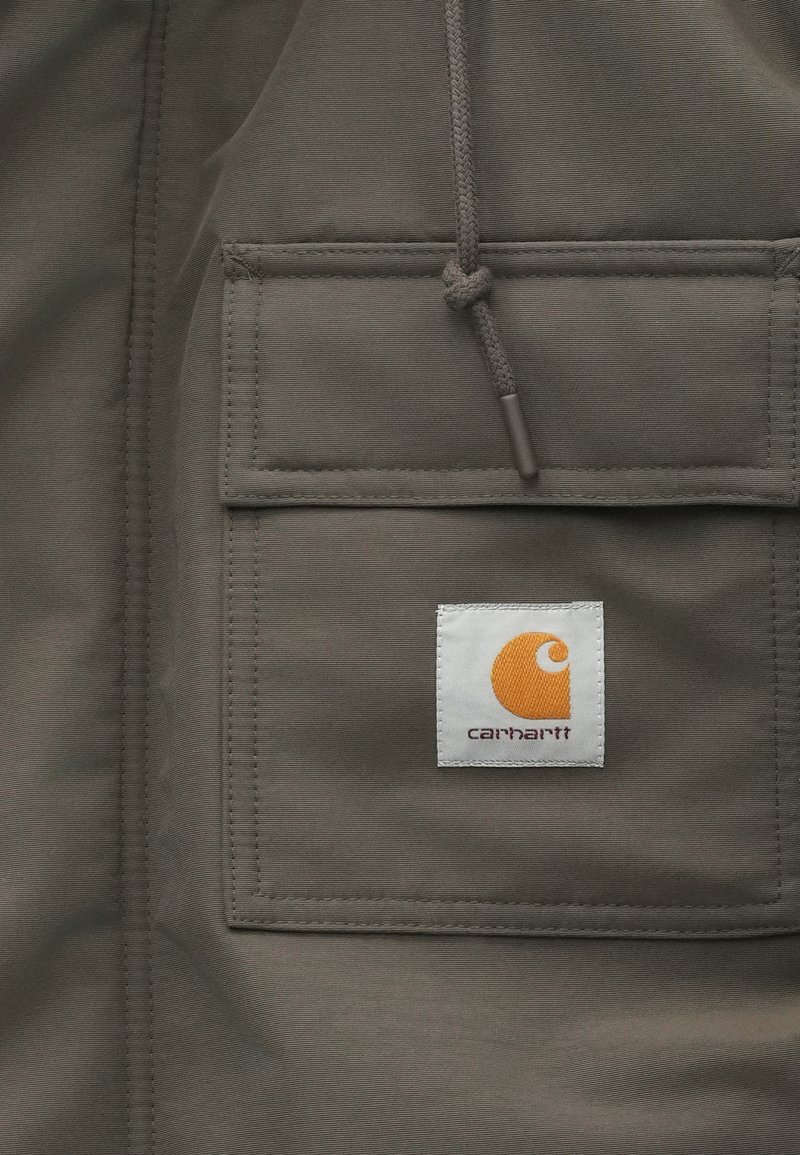 Giacca in tessuto grigio con una tasca frontale e un logo quadrato bianco. Il logo include un vortice arancione e il testo "carhartt" qui sotto.