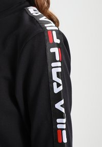 Pull-over noir avec un tissu texturé et un panneau brillant noir sur l'épaule affichant "FILA" en lettres blanches et rouges audacieuses.