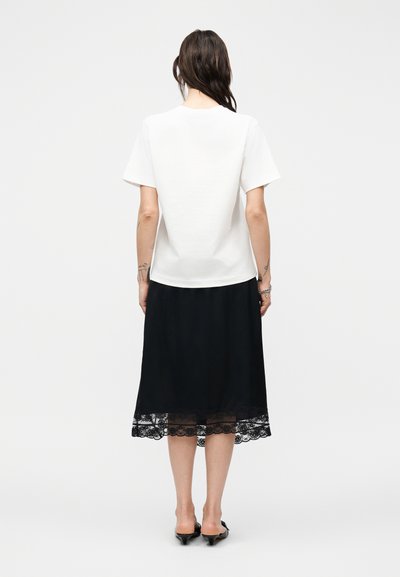 Γυναίκα που στέκεται γυρισμένη με την πλάτη, φορώντας λευκό μπλουζάκι με κοντά μανίκια, μαύρη midi φούστα με δαντέλα στο τελείωμα και μαύρα παπούτσια με τακούνι kitten, πάνω σε γκρίζο φόντο.