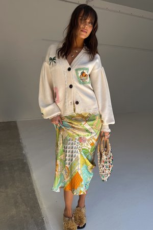 Cardigan blanc surdimensionné avec des patches brodés, jupe portefeuille à motifs dans des teintes de vert et de jaune, et slides beige moelleux. Sac en osier.