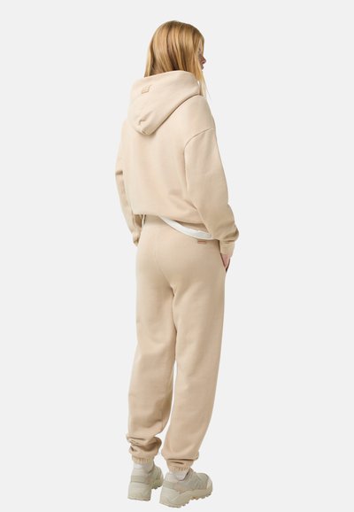 Lys beige hoodie og joggers lavet af blødt stof, med en afslappet pasform, ribbede manchetter og et diskret logo nær taljen.