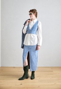 Chaleco de mezclilla con botones sobre una camisa blanca, combinado con una falda de mezclilla con una abertura frontal. Botas altas de ante verde.