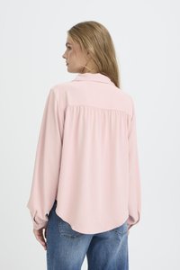 Donna bionda che indossa una blusa lunga a maniche lunghe rosa chiaro e jeans blu, di profilo con uno sfondo bianco neutro.