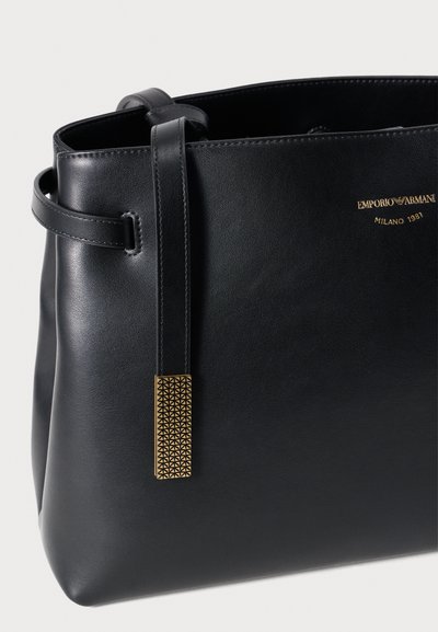 Μαύρη δερμάτινη τσάντα tote με δομημένο σχήμα, με σφιχτά ραμμένα άκρα, μια ετικέτα με χρυσές λεπτομέρειες και επάνω χερούλι για μεταφορά.