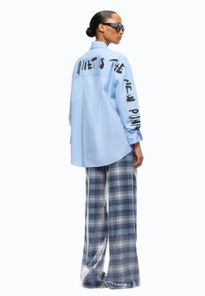 Mujer con camisa azul claro de gran tamaño con texto "Quiet is the new punk" y pantalones anchos de cuadros, posando de lado con gafas de sol.