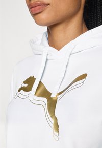 Haut à capuche blanc avec un grand logo Puma doré entouré d'un contour noir. Le tissu semble doux avec un aspect texturé. Les cordons sont visibles.