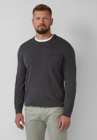 Donkergrijze lange mouwen trui met ronde hals, gemaakt van zacht materiaal met geribbelde manchetten. Bevat een klein logo op de linkerborst. Gedragen over een wit shirt.