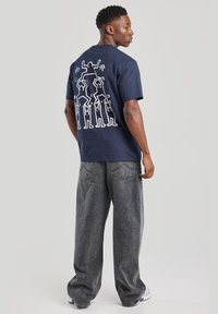 Camiseta azul marino con diseño gráfico blanco de figuras estilizadas en la parte de atrás. Corte relajado, mangas cortas y material de algodón. Combinada con jeans anchos grises.