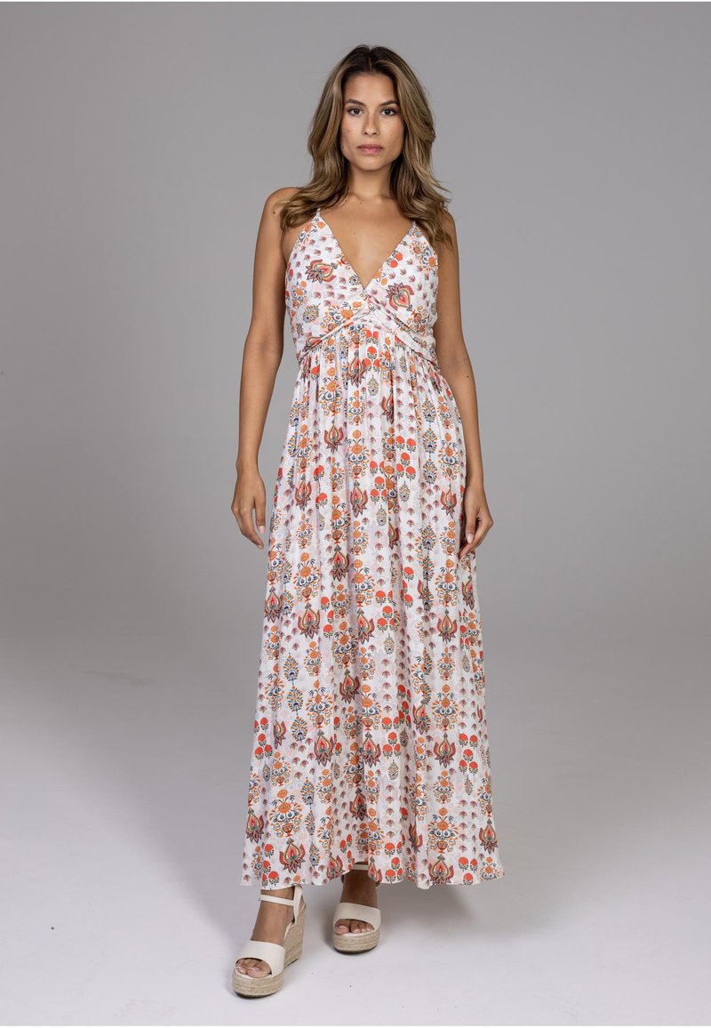 Femme debout portant une robe maxi blanche sans manches avec des motifs floraux orange et rouge et des sandales compensées beige sur fond gris.