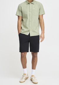 Chemise vert clair à manches courtes avec une finition texturée, boutons sur le devant et une poche. Portée avec un short noir et des baskets beiges.