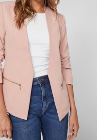 Blazer rose blush avec ouverture devant, manches trois-quarts et deux poches zippées. Associé à un haut blanc et un jean bleu taille haute.