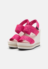 Růžové platformové sandály z látky se širokým páskem s logem "Calvin Klein Jeans". Zvýšená bílá wedge podrážka s černým gripem.