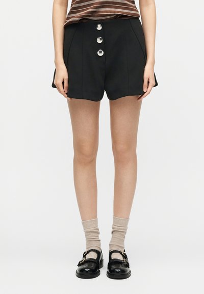 Γυναίκα που φοράει μαύρα σορτς με ψηλή μέση και τρία μεγάλα μπροστινά κουμπιά, μπεζ κάλτσες και μαύρα παπούτσια στιλ Mary Jane.
