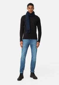 Schwarzer Pullover, dunkelblauer Schal, hellblaue Jeans und schwarze Stiefel. Der Schal hat Fransen; die Jeans sind eng geschnitten mit einer verwaschenen Textur.