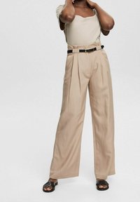 Femme portant un pantalon beige taille haute à jambes larges avec ceinture noire et haut beige, debout, mains près du cou, sandales noires aux pieds.