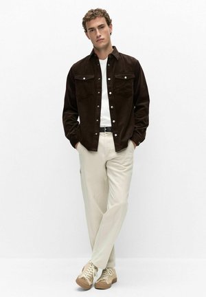 Homme debout, les mains dans les poches, portant une chemise marron foncé boutonnée, un t-shirt blanc, un pantalon beige clair et des baskets beiges.