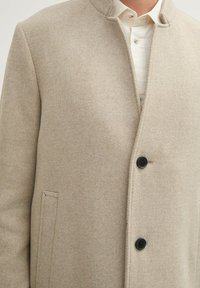 Beige Wollmantel mit strukturierter Oberfläche, mit hohem Kragen, zwei schwarzen Knöpfen und Seitentaschen. Maßgeschneiderter Schnitt, klare Linien.