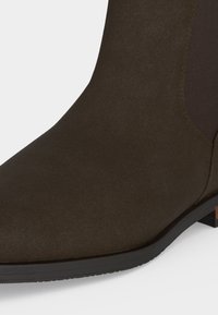 Gros plan d’une botte cheville en daim marron foncé avec un panneau latéral élastique et une semelle en caoutchouc noire.