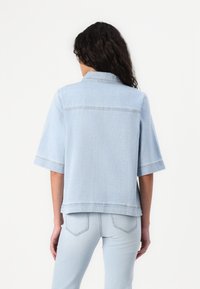Jaqueta de ganga azul claro com mangas curtas, colarinho e detalhes cosidos. Apresenta uma bainha reta e um corte descontraído.