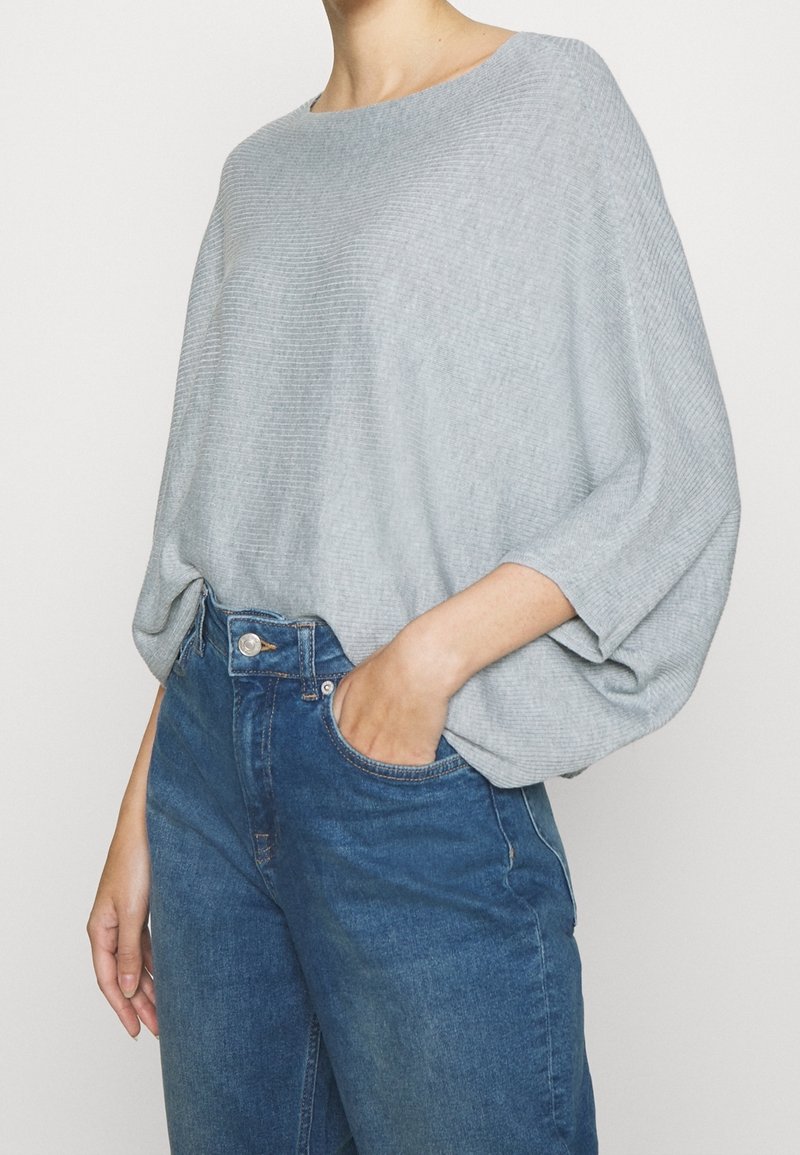 Personne portant un pull à côtes ample gris clair et un jean bleu taille haute, une main dans la poche, sur un fond uni.