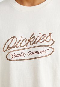 Kremo spalvos medvilninė marškinė su rudo virvės stiliaus spauda, kuriame užrašyta "Dickies" ir "Quality Garments" stilizuotu šriftu.