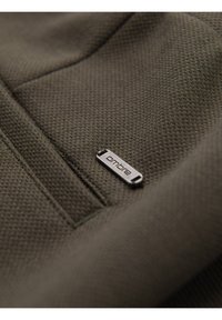 Gros plan d’un tissu texturé olive foncé avec une petite étiquette métallique argentée marquée "ombre", cousue près d’une couture.