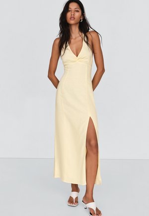 Kvinde med langt mørkt hår iført en bleg gul ærmeløs kjole med slids op til låret og hvide hælede sandaler.