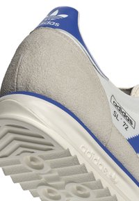 Tenisky s šedým semišovým svrškem, modrými akcenty, bílou mezipodešví a texturovanou gumovou podrážkou s výrazným logem Adidas a modelem číslo.