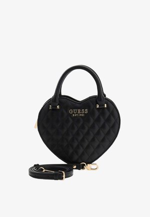 Schwarze, gesteppte, herzförmige Handtasche mit zwei Griffen, goldfarbenen Beschlägen, "GUESS EST 1981" Logo und abnehmbarem Schulterriemen, der zusammengerollt danebenliegt.